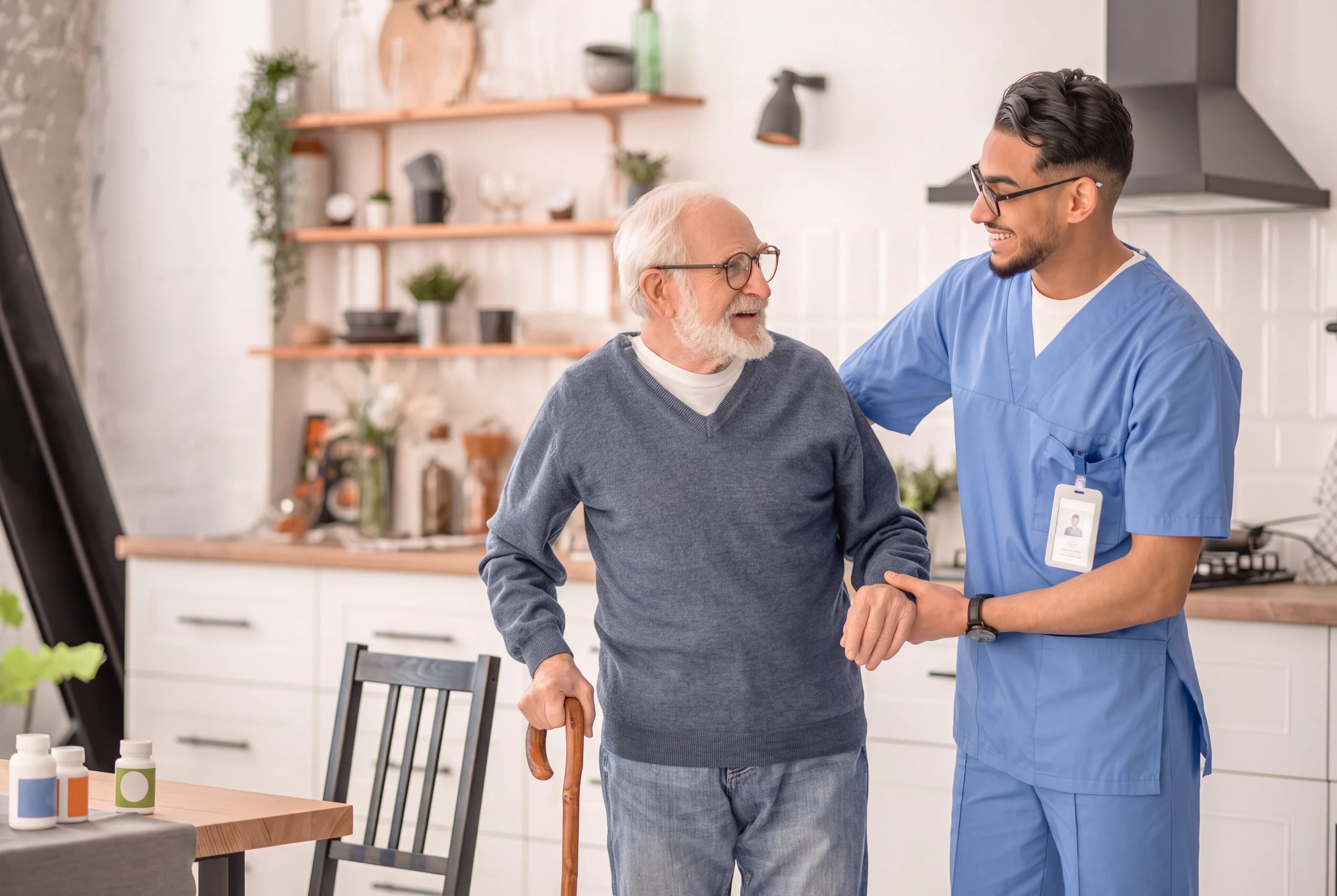 A home health caregiver supporting an older adult with a walking cane in his kitchen